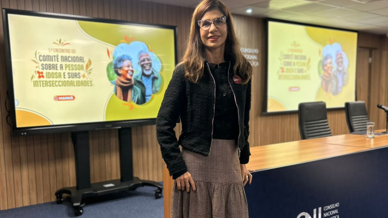 imagem mostra a juíza, em pé, sorrindo para a câmera, tendo ao fundo painel eletrônico com a identidade visual do evento (imagem nas cores verde, amarelo e laranja, com foto de um casal negro idoso e o título "1º Encontro do Comitê Nacional sobre a Pessoa Idosa e suas Intersecccionalidades"