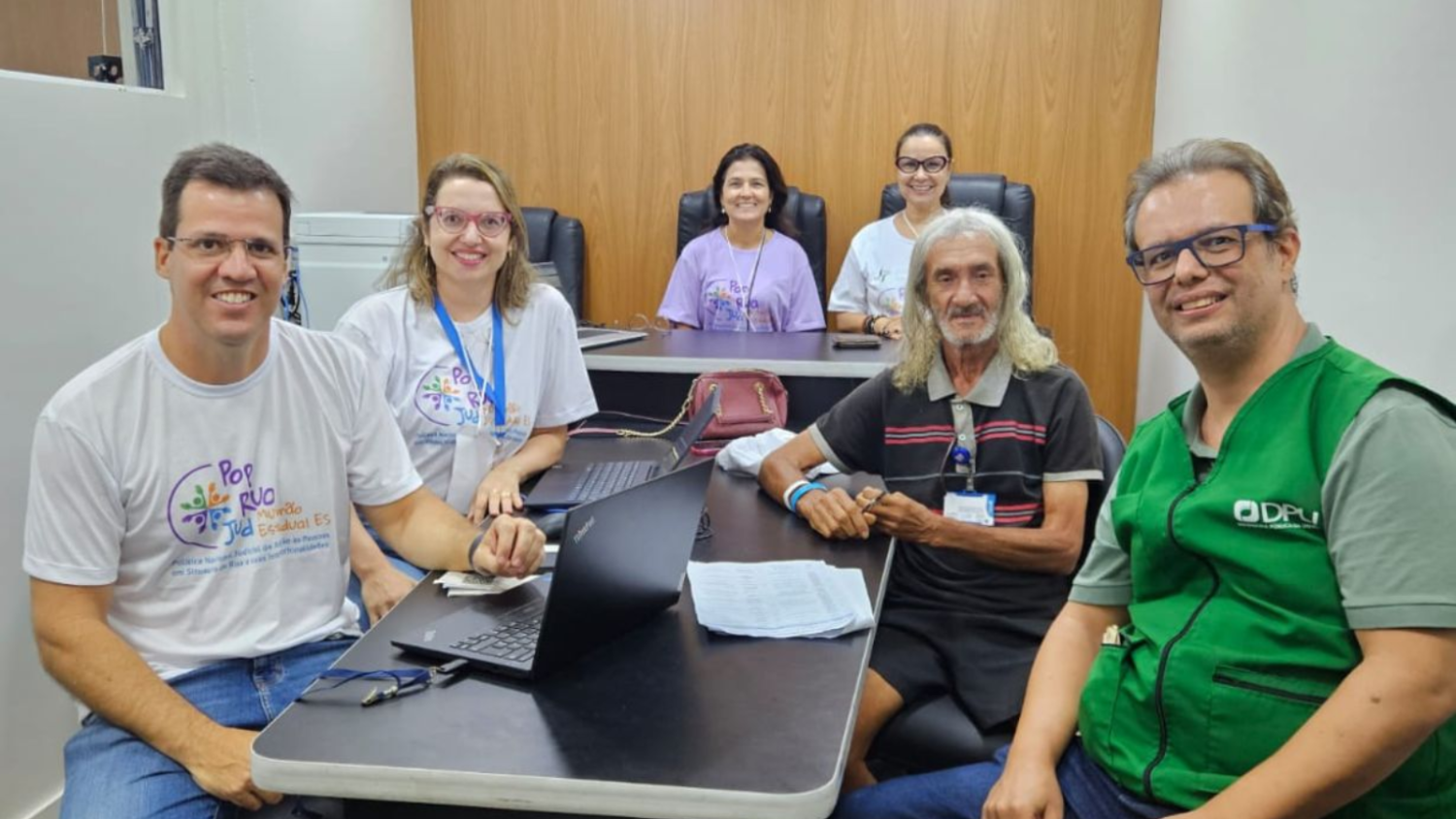 Grupo de voluntários do Mutirão PopRuaJud atende um homem em uma sala, sentados ao redor de uma mesa com notebooks, todos olhando para a câmera.
