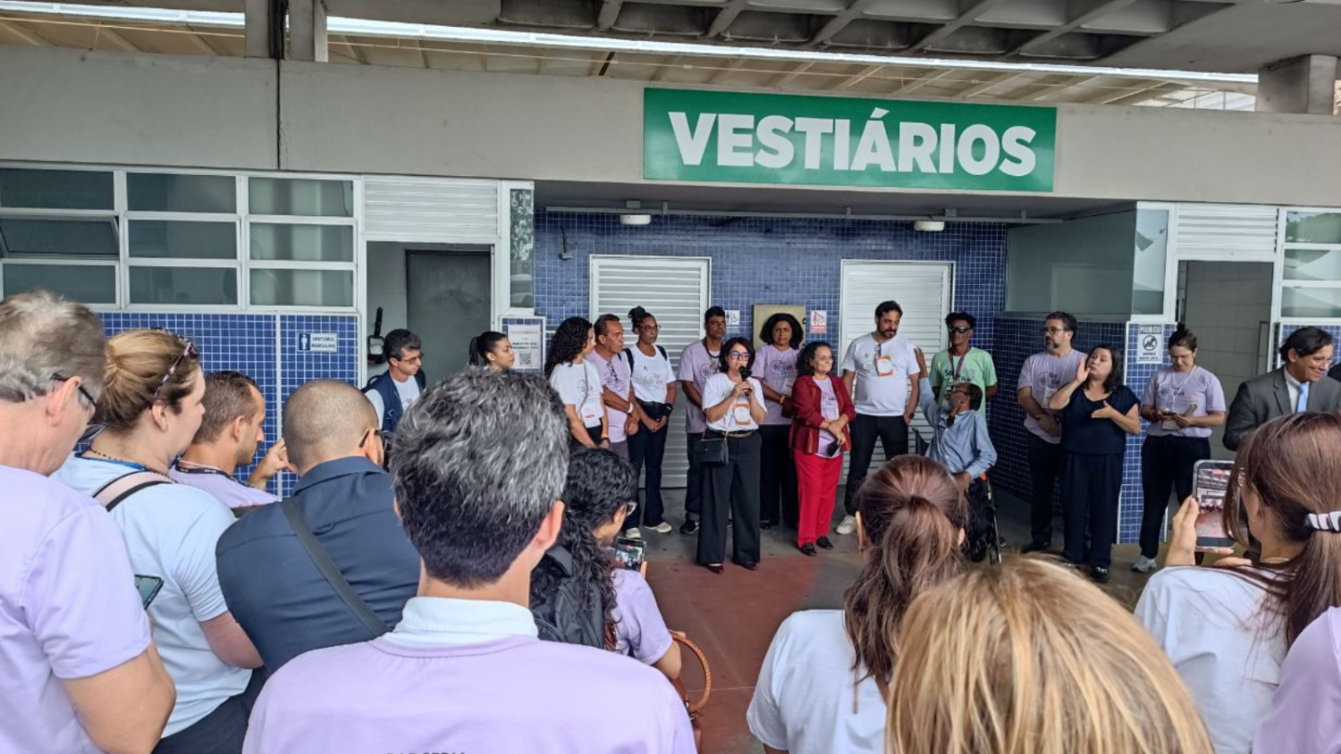 Grupo de pessoas reunidas em frente a uma área de vestiários durante fala de abertura do Mutirão PopRuaJud; uma mulher ao centro usa microfone enquanto voluntários e participantes acompanham.