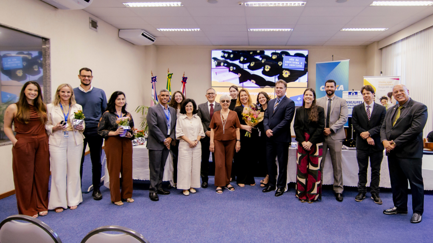 Encerramento da cerimônia com servidores e autoridades reunidos, uma servidora ao centro segura um buquê de flores.