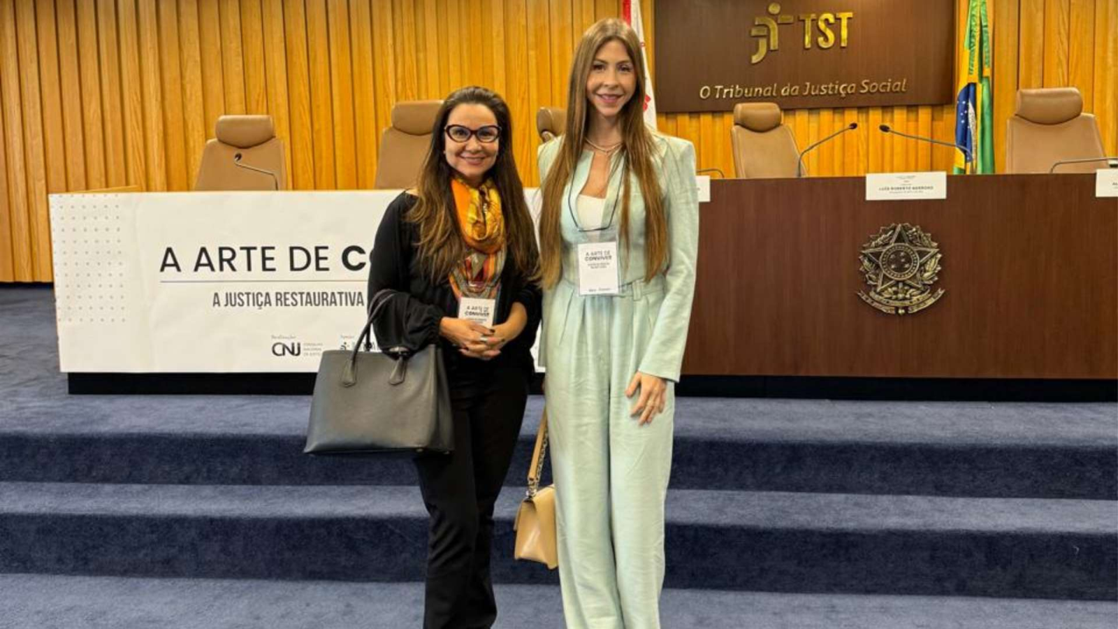 Duas mulheres de pé, posando para a câmera. Ao fundo, mesa de madeira escura com brasão da República em bronze