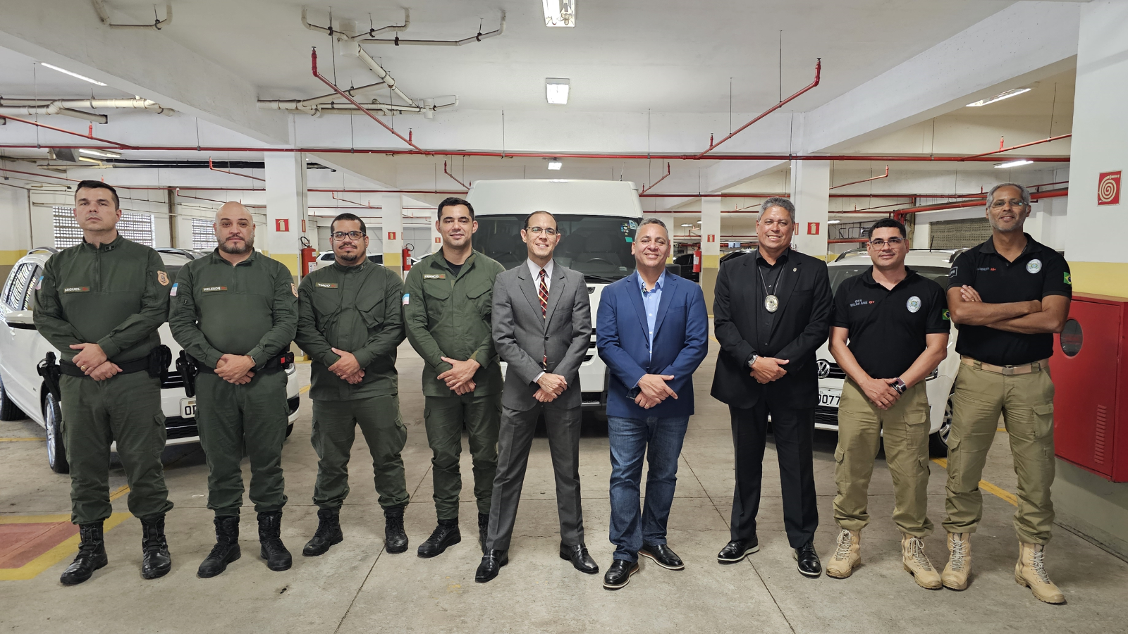 9 homens, sendo quatro de uniforme verde militar, dois de terno, um de calça e blazer pretos e dois de calça cáqui e camisa polo preta