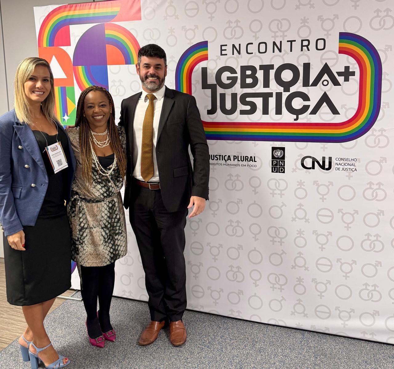 A imagem mostra três magistrados da Justiça Federal do Rio de Janeiro posando lado a lado para uma foto em ambiente interno e institucional. Ao centro está a juíza federal Adriana Cruz, que também é secretária-geral do Conselho Nacional de Justiça (CNJ). À direita dela, na imagem, está o juiz federal Marcel da Silva Augusto Corrêa, convocado em auxílio à Presidência do CNJ. À esquerda, aparece a juíza federal Giovana Brantes Calmon. Todos estão sorrindo e vestidos formalmente, com roupas sociais. Ao fundo, há uma parede clara e parte de uma bandeira nacional visível, reforçando o caráter oficial do encontro.