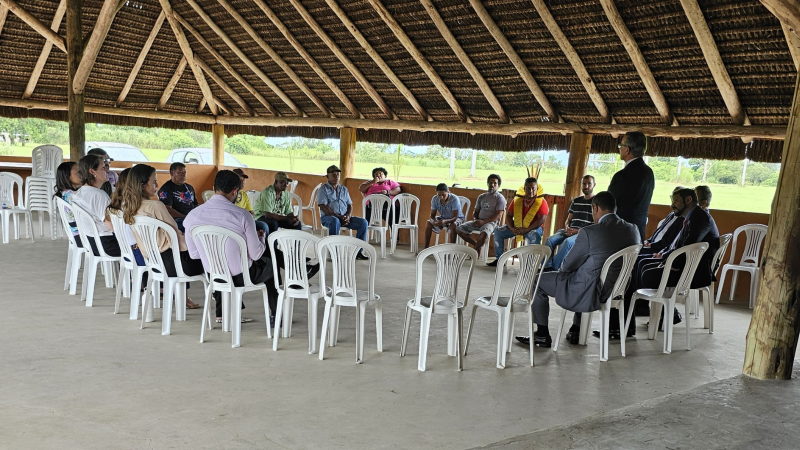 Homens e mulheres sentados em círculo, em cadeiras plásticas brancas, sob um telhado de eucalipto e palha. Um deles fala de pé (presidente do TRE)