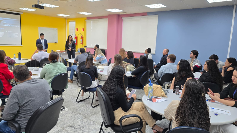 Sala de aula, com aproximadamente 40 pessoas sentadas, divididas em grupos. À frente, o juiz e a juíza dando aula