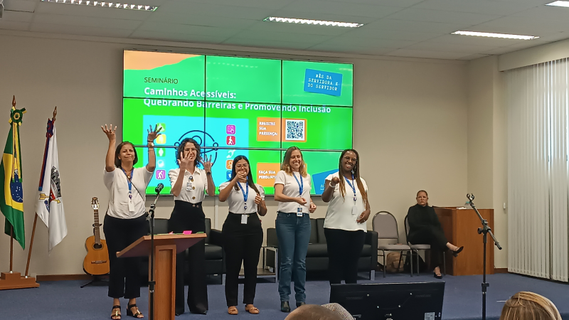 Cinco mulheres, vestindo camisetas brancas e crachás, fazem uma apresentação em Libras no auditório da Justiça Federal. Ao fundo, o telão exibe o tema “Caminhos Acessíveis: Quebrando Barreiras e Promovendo Inclusão”, e uma intérprete de Libras está sentada.