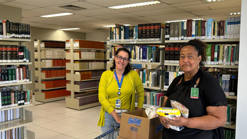 Uma mulher de roupa amarela entrega donativos a outra mulher de roupa preta. Ao fundo, estantes com livros
