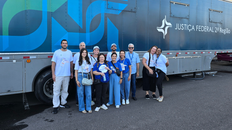 Grupo de servidores e participantes posa para foto em frente a um caminhão azul com a identificação da Justiça Federal. A maioria veste camisetas azuis do evento e crachás.