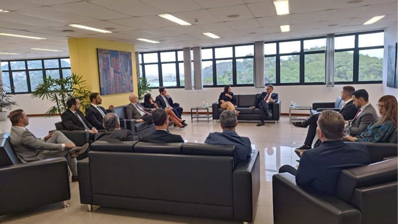 Sala ampla de reunião com grandes janelas ao fundo, mostrando área verde. Cerca de 15 pessoas, em sua maioria homens de terno, estão sentadas em sofás pretos dispostos em círculo, participando de uma conversa institucional.