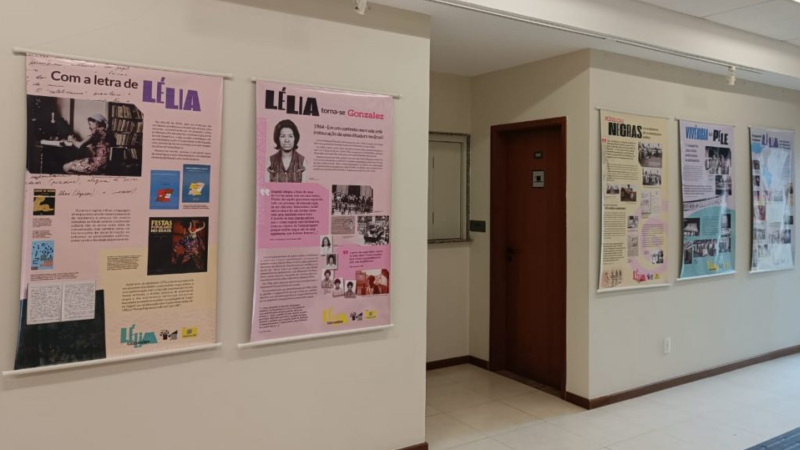#PraTodosVerem: Fotografia de um corredor interno da Justiça Federal com banners da exposição sobre Lélia Gonzalez afixados nas paredes. Os painéis apresentam textos, fotografias e documentos sobre a trajetória da intelectual, professora e ativista. Ao fundo, há uma porta de madeira e a circulação do espaço expositivo.