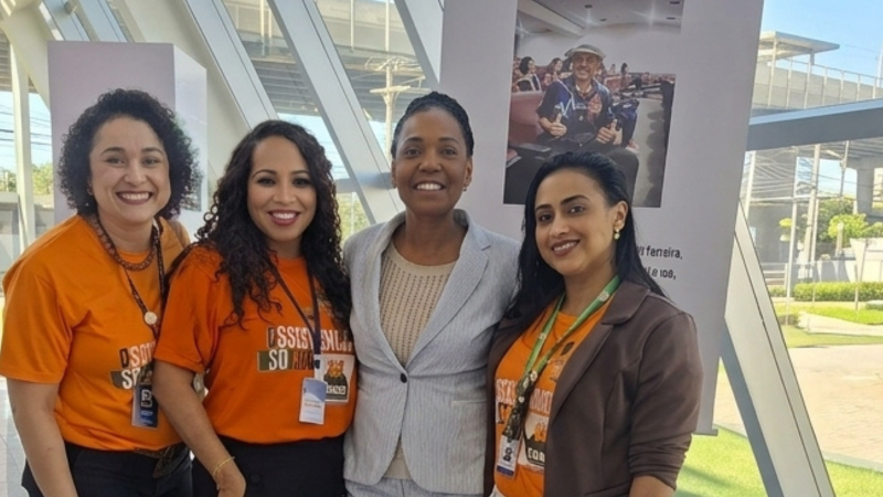 Quatro mulheres posam sorrindo para a câmera, sendo três mulheres brancas ou pardas de camisas laranja e, ao centro, uma mulher negra de terninho listrado azul e branco