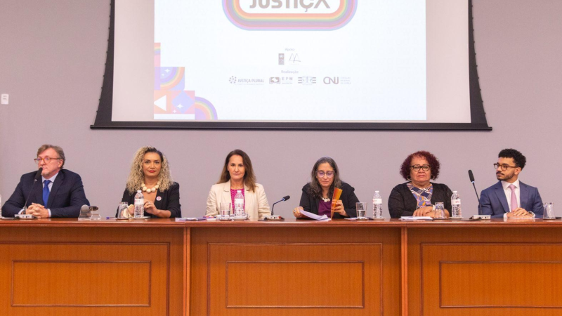 Mesa diretora de madeira com dois homens e quatro mulheres. Ao fundo, telão com logotipos do evento 