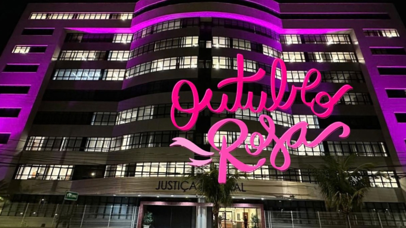 Fachada da sede da Justiça Federal, em Vitória, com iluminação na cor rosa. Sobre a foto, o texto "Outubro Rosa", em letras também cor-de-rosa