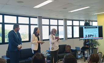 O professor Gustavo Forde está em pé, à frente da sala, segurando um livro e apresentando-o ao público. Ao lado dele, uma mulher segura outros exemplares. Um homem de terno observa. Ao fundo, uma tela exibe slide com texto e imagens. Participantes assistem sentados.