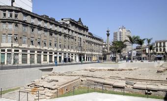 Vista ampla de uma praça urbana com estruturas arqueológicas de pedra escavadas em primeiro plano, cercadas por guarda-corpos metálicos. Ao fundo, há edifícios históricos de vários andares, palmeiras e um monumento em coluna no centro do espaço público. O céu está limpo e azul, destacando o contraste entre as ruínas preservadas e a paisagem urbana do centro da cidade.