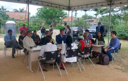 audiência com seis homens e três mulheres sentados à mesa, sob uma cobertura de lona