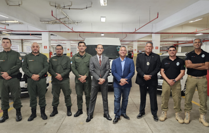 9 homens de pé, de frente para a câmera. Ao fundo, três carros brancos