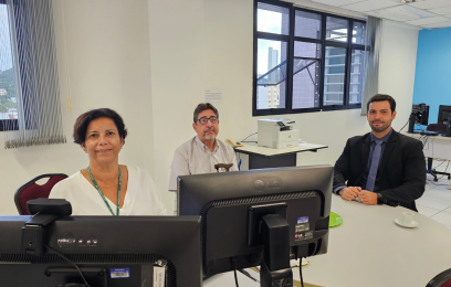 Uma mulher e dois homens em volta de uma mesa. Ela, ao computador