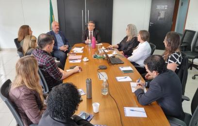 Um grupo de pessoas está reunido ao redor de uma mesa retangular de madeira em uma sala de reuniões. Sobre a mesa há diversos itens: cadernos, canetas, copos, uma garrafa de água, um notebook, um recipiente de vidro com água, cabos e uma garrafa térmica rosa. A sala tem paredes brancas e piso de cerâmica clara. Ao fundo, há um armário preto com duas portas, e ao lado dele, uma bandeira do Brasil. As pessoas estão envolvidas em uma conversa, algumas tomando notas e outras ouvindo com atenção.
