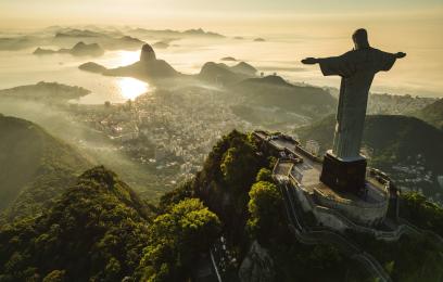 Descrição da imagem: A imagem apresenta uma vista aérea do Cristo Redentor, icônica estátua localizada no topo do Morro do Corcovado, no Rio de Janeiro, Brasil. A estátua, com os braços abertos, transmite uma mensagem de paz e acolhimento. Ao fundo, vê-se a cidade do Rio de Janeiro, com destaque para a Baía de Guanabara e o Pão de Açúcar, ambos marcos naturais emblemáticos. A paisagem combina elementos urbanos e naturais, revelando a integração entre a cidade e seu entorno montanhoso e costeiro.