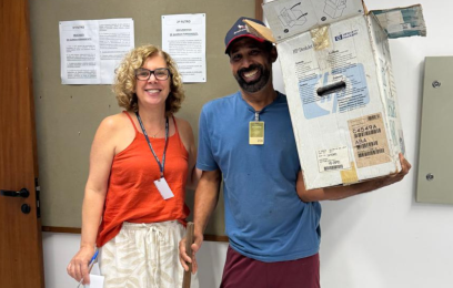 Uma mulher e um homem sorrindo para a câmera. Ele segura uma caixa média de papelão, com materiais dentro