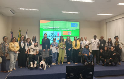Grupo diverso de pessoas posa para foto no auditório da Justiça Federal do Espírito Santo, em frente a um telão com o tema “Caminhos Acessíveis: Quebrando Barreiras e Promovendo Inclusão”. Entre os participantes estão pessoas com deficiência, servidoras e servidores, além de uma cadela de apoio emocional