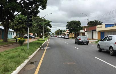Fotografia de uma avenida urbana em um dia nublado. A via é larga, com duas faixas de rolamento em cada sentido, separadas por uma linha central descontínua. O asfalto está escuro e ligeiramente úmido, com alguns pequenos remendos visíveis.  À esquerda, há uma calçada com grama bem cuidada, árvores de médio porte e um semáforo apagado ou inativo. Algumas casas térreas aparecem recuadas, com jardins frontais. À direita, há mais construções baixas, incluindo estabelecimentos comerciais simples.