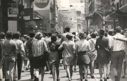 Fotografia em preto e branco de uma rua movimentada no centro urbano, com muitas pessoas caminhando juntas, vistas de costas. Há prédios altos, placas comerciais e uma multidão ocupando a via, sugerindo uma manifestação