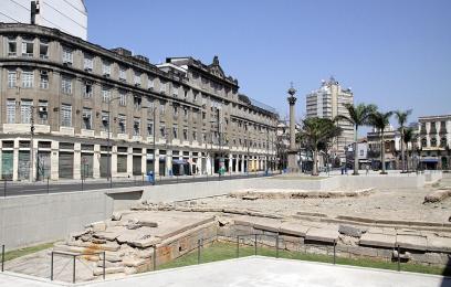Vista ampla de uma praça urbana com estruturas arqueológicas de pedra escavadas em primeiro plano, cercadas por guarda-corpos metálicos. Ao fundo, há edifícios históricos de vários andares, palmeiras e um monumento em coluna no centro do espaço público. O céu está limpo e azul, destacando o contraste entre as ruínas preservadas e a paisagem urbana do centro da cidade.