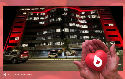 Fotografia noturna do edifício-sede da JFES iluminado em vermelho, em referência à campanha Junho Vermelho. No canto inferior direito da imagem, mãos em concha seguram um coração vermelho com o símbolo de uma gota de sangue e uma cruz branca