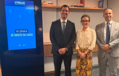 Foto de três magistrados em pé, lado a lado, em ambiente interno. À esquerda, um juiz veste terno escuro e gravata; ao centro, uma juíza usa camisa clara e saia estampada; à direita, outro juiz veste terno cinza claro e gravata. Ao lado deles, há um painel eletrônico azul com a inscrição “VIII Jornada de Direito da Saúde”